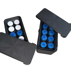 May include: Two black rectangular storage cases with textured lids, each containing multiple small glass vials. One case holds vials with white caps, while the other case contains vials with blue caps, arranged in two parallel rows inside the containers.