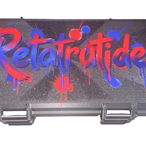 May include: A rectangular black hard-shell carrying case featuring a textured surface. The top of the case displays the word Retatrutide in a stylized, dripping graffiti font with vibrant red and blue paint splatter accents.