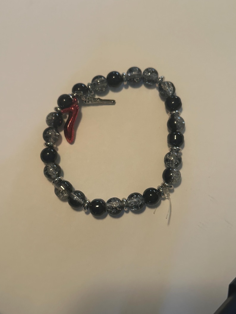 May include: A beaded bracelet featuring black and clear crackle beads, interspersed with silver-toned spacers. A small red horn charm and a silver-toned charm are attached. The bracelet is designed to be worn around the wrist.