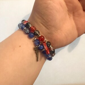 May include: A beaded bracelet set featuring blue, red, black, and clear beads. One bracelet has a silver cross charm and a red charm. The bracelets are worn on a wrist.