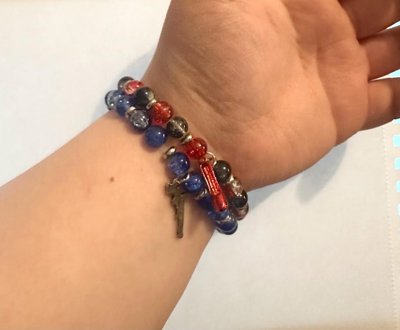 May include: A beaded bracelet set featuring blue, red, black, and clear beads. One bracelet has a silver cross charm and a red charm. The bracelets are worn on a wrist.