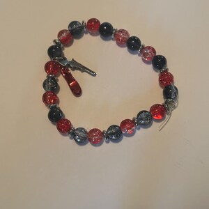 May include: A beaded bracelet featuring red, black, and gray beads with silver accents. The bracelet includes a small silver gun charm and a red charm. The beads are strung on a clear elastic cord.