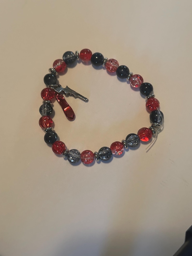 May include: A beaded bracelet featuring red, black, and gray beads with silver accents. The bracelet includes a small silver gun charm and a red charm. The beads are strung on a clear elastic cord.
