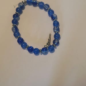 May include: A blue beaded bracelet with a silver-colored charm shaped like a gun. The bracelet features round, crackled blue beads and silver-colored spacers. The bracelet is on a white surface.
