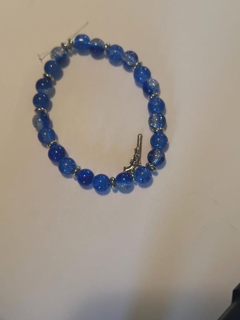 May include: A blue beaded bracelet with a silver-colored charm shaped like a gun. The bracelet features round, crackled blue beads and silver-colored spacers. The bracelet is on a white surface.