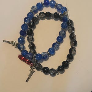 May include: Two beaded bracelets with blue, black, and silver beads. One bracelet has a silver charm of a gun, and the other has a red charm of a high heel shoe and a silver charm of a sword. The bracelets are on a white surface.