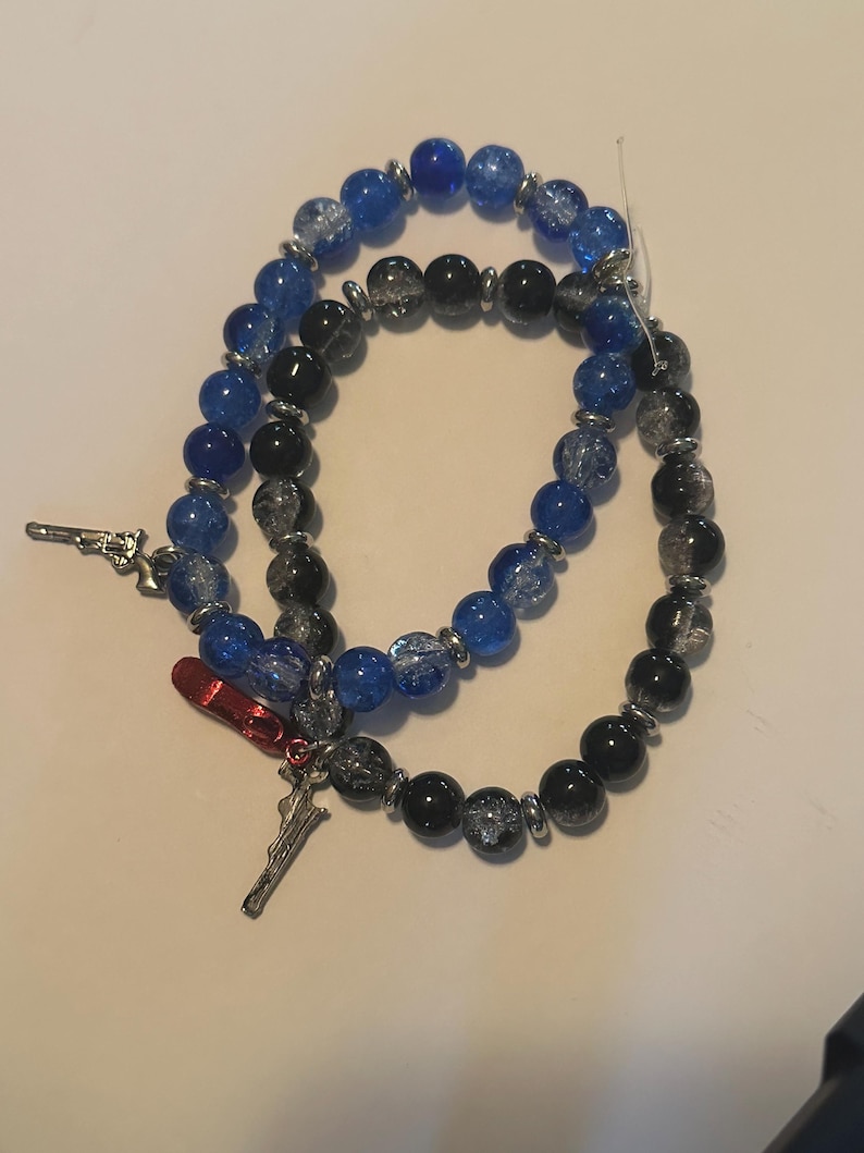 May include: Two beaded bracelets with blue, black, and silver beads. One bracelet has a silver charm of a gun, and the other has a red charm of a high heel shoe and a silver charm of a sword. The bracelets are on a white surface.