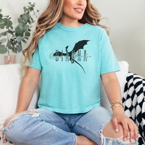 Puede incluir: Camiseta azul claro con un dise&ntilde;o de drag&oacute;n negro y el texto "Queen of Dragons". La camiseta es de tejido suave y tiene un corte relajado. El dise&ntilde;o est&aacute; centrado en la parte delantera.