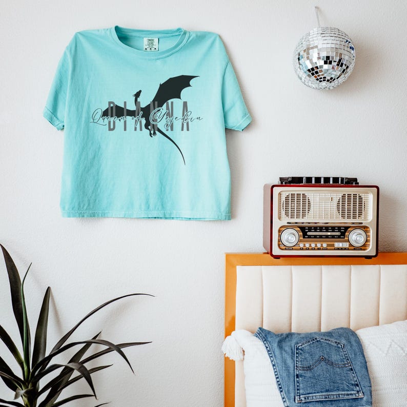 Puede incluir: Camiseta azul claro con un dise&ntilde;o de drag&oacute;n negro y el texto "Queen of Yggdrasil". La camiseta est&aacute; colgada en una pared blanca, con una bola de discoteca y una radio vintage en el fondo.