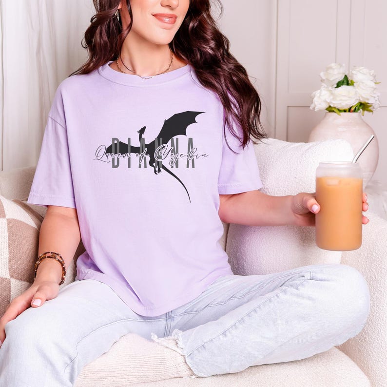 Puede incluir: Camiseta lila claro con un dise&ntilde;o de drag&oacute;n negro y el texto "Queens of Targaryen". La camiseta tiene mangas cortas. La persona sostiene un vaso de caf&eacute; helado. La camiseta es de material suave.