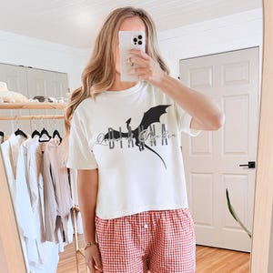 Puede incluir: Camiseta color crema con un gráfico de dragón negro y el texto "Queen of Eyebin". La camiseta se combina con pantalones cortos de pijama a cuadros rojos y blancos. La imagen se toma frente a un espejo.
