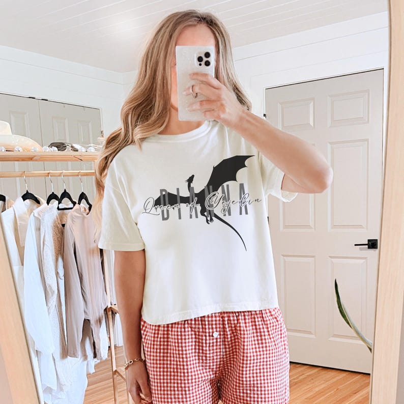 Puede incluir: Camiseta color crema con un gr&aacute;fico de drag&oacute;n negro y el texto "Queen of Eyebin". La camiseta se combina con pantalones cortos de pijama a cuadros rojos y blancos. La imagen se toma frente a un espejo.
