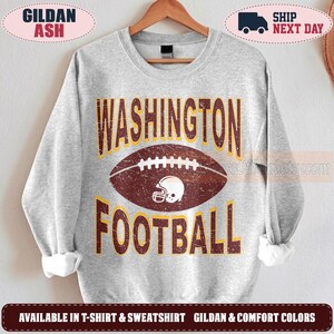 May include: A light gray crewneck sweatshirt featuring a distressed graphic of a brown football and a white helmet. The text WASHINGTON FOOTBALL is printed in large, bold, dark red letters with a yellow outline above and below the football.