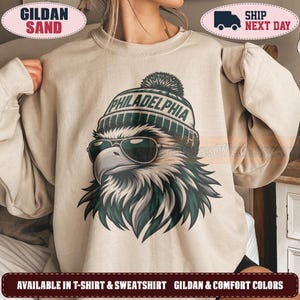 Könnte beinhalten: Ein sandfarbenes Sweatshirt mit Rundhalsausschnitt und einer Grafik eines Adlers, der eine dunkle Sonnenbrille und eine Strickmütze mit Bommel trägt. Auf der Mütze steht in dunkelgrünen Buchstaben PHILADELPHIA. Der Adler hat weiße und dunkelgrüne Federn.