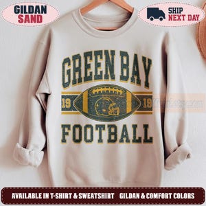 Könnte beinhalten: Ein sandfarbenes Sweatshirt mit Rundhalsausschnitt und einem abgenutzten Grafikdruck. Das Design zeigt den Text GREEN BAY FOOTBALL in Dunkelgrün mit gelben Umrandungen, zentriert um eine Football-Illustration mit einem Helm-Symbol und der Zahl 19.