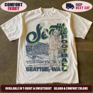 May include: An ivory short-sleeve t-shirt featuring a graphic print of a football player in a jersey with the number 14, a stadium, and the text Seattle Football Est. 1976 Seattle, WA in dark blue and green ink on the front of the garment.