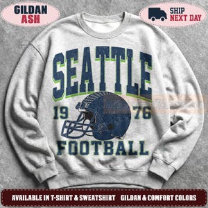 May include: A light grey crewneck sweatshirt featuring a distressed graphic print. The design includes the text SEATTLE in large dark blue letters with green outlines, the number 1976, and the word FOOTBALL above a dark blue football helmet.