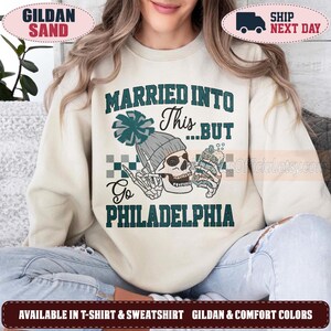 Puede incluir: Una sudadera de cuello redondo color arena con un gráfico de un esqueleto que lleva un gorro gris y sostiene una bebida. El texto dice "MARRIED INTO This...BUT Go PHILADELPHIA" en verde oscuro, con un patrón de cuadros tras el esqueleto.