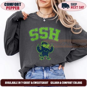May include: A charcoal gray long-sleeve sweatshirt featuring the text SSH in large, distressed lime green block letters. Below the text is a graphic of a dark blue bird wearing lime green sunglasses and gesturing with both middle fingers.
