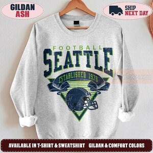 May include: A light gray crewneck sweatshirt featuring a distressed graphic print. The design includes the text FOOTBALL SEATTLE ESTABLISHED 1976 in dark blue and green, centered above a dark blue football helmet and a green triangular background.