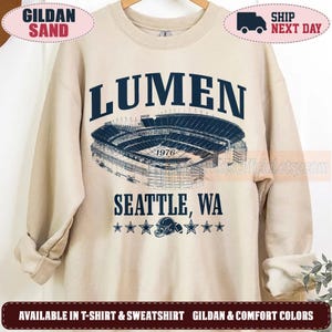 May include: A sand-colored crewneck sweatshirt featuring dark blue text that reads LUMEN and SEATTLE, WA. A detailed line-art illustration of a stadium is centered between the text, with the year 1976, a football helmet, and six stars below.