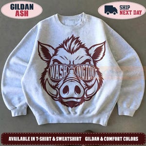 May include: A light gray crewneck sweatshirt featuring a dark maroon graphic of a wild boar wearing sunglasses. The lenses of the sunglasses contain the text WASH and INGTON respectively. The sweatshirt has long sleeves and a ribbed collar.