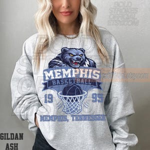 May include: A light gray sweatshirt featuring a blue and white Memphis Basketball design. The design includes a bear graphic, basketball, and the text "MEMPHIS BASKETBALL 1995 MEMPHIS, TENNESSEE". The sweatshirt is made of Gildan Ash material.