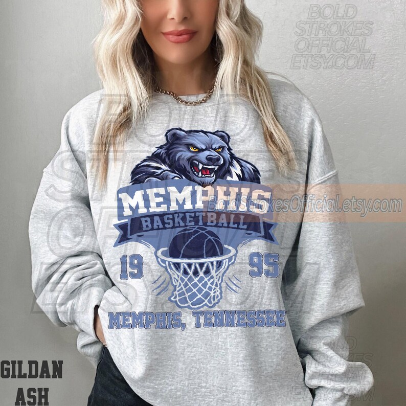 May include: A light gray sweatshirt featuring a blue and white Memphis Basketball design. The design includes a bear graphic, basketball, and the text "MEMPHIS BASKETBALL 1995 MEMPHIS, TENNESSEE". The sweatshirt is made of Gildan Ash material.