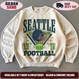 May include: A sand-colored crewneck sweatshirt featuring a dark blue and green graphic print. The design includes the text SEATTLE FOOTBALL, the year 1976, a football helmet, and a football field illustration with stars on a light background.