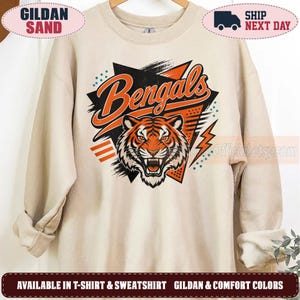 May include: A sand-colored crewneck sweatshirt featuring a graphic print of a tiger face with the word Bengals in orange and black lettering. The design includes geometric shapes, orange lightning bolts, and blue dots on a light beige fabric background.