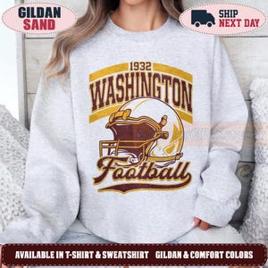 May include: A light gray crewneck sweatshirt featuring a graphic print of a football helmet in yellow and maroon. The text reads 1932 WASHINGTON Football in a bold, arched font. The design is centered on the chest of the long-sleeved garment.