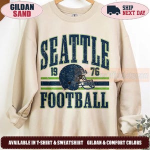 May include: A sand-colored crewneck sweatshirt featuring a distressed navy blue and green graphic. The text reads SEATTLE 1976 FOOTBALL, centered around a navy blue football helmet illustration. The design includes horizontal stripes in navy and green.