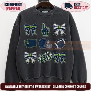 May include: A charcoal grey crewneck sweatshirt featuring a grid of nine graphic icons in navy, lime green, and white. The designs include four bows, a number one foam finger, a map of Washington state, a football helmet, a football, and pom-poms.