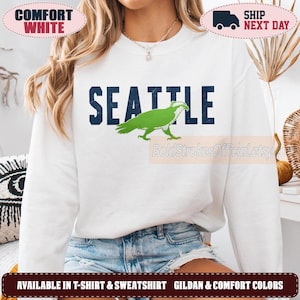 May include: A white long-sleeve crewneck sweatshirt featuring the word SEATTLE in large dark blue block letters. A bright green graphic of an osprey bird is positioned over the letters. The design has a slightly distressed, textured appearance.