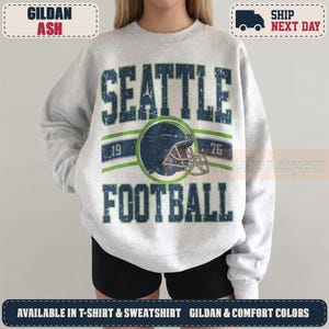 May include: A light gray crewneck sweatshirt featuring a distressed dark blue and green graphic. The design includes the text SEATTLE FOOTBALL, the numbers 19 and 76, and a central dark blue football helmet illustration with a white face mask.
