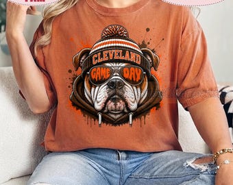 Puede incluir: Una camiseta de manga corta color óxido con el gráfico de un bulldog que lleva una sudadera marrón, un gorro a rayas y gafas de sol. El texto CLEVELAND está impreso en el gorro y GAME DAY es visible en los cristales de las gafas.