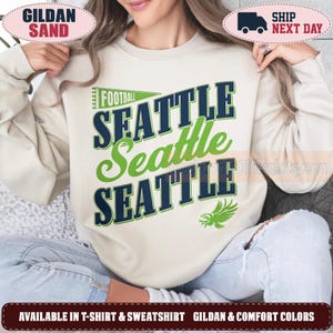 May include: A sand-colored crewneck sweatshirt featuring a graphic print with the word SEATTLE repeated three times in alternating navy blue and lime green block lettering, a green football pennant, and a small green bird icon on the front.