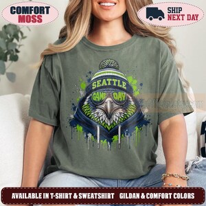 May include: A moss green short-sleeve t-shirt featuring a graphic of an eagle wearing a blue and green knit beanie and sunglasses. The text "SEATTLE" and "GAME DAY" is printed on the beanie and sunglasses in a bold, green font.