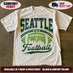 May include: An ivory short-sleeve t-shirt featuring a graphic print with the text SEATTLE 1976 Football. The design includes a green football shape containing a white city skyline silhouette, with dark blue and green lettering and outlines.