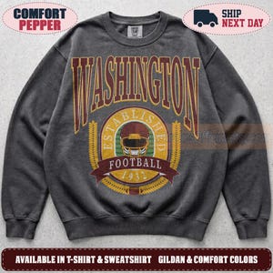 May include: A charcoal grey crewneck sweatshirt featuring a large graphic on the chest. The text reads WASHINGTON in bold letters above a circular emblem with the words ESTABLISHED FOOTBALL 1932 and a central illustration of a football helmet.