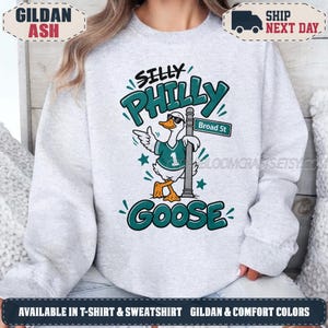 Könnte beinhalten: Ein hellgraues Sweatshirt mit Rundhalsausschnitt und einer Grafik einer Gans, die eine Sonnenbrille und ein blaugrünes Trikot mit der Nummer eins trägt. Die Gans lehnt an einem Straßenschild mit der Aufschrift Broad St. Der Text lautet Silly Philly Goose.