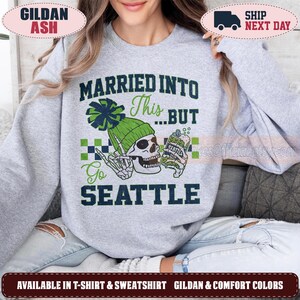 May include: A light gray crewneck sweatshirt featuring a graphic of a skull wearing a green beanie and holding a drink. The text reads MARRIED INTO This...BUT Go SEATTLE. The design includes a green and navy blue checkered pattern element.