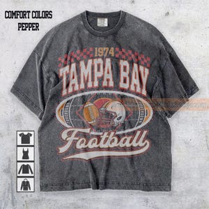 Puede incluir: Camiseta gris oscuro con un diseño de fútbol vintage. La camiseta presenta el texto "TAMPA BAY Football 1974" en rojo y blanco, junto con un gráfico de fútbol y casco. La camiseta es de color pimienta.