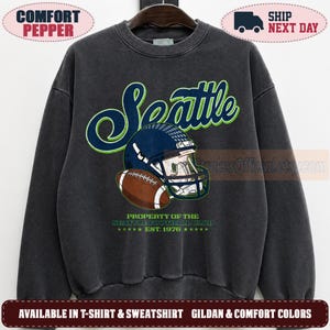 May include: A charcoal grey crewneck sweatshirt featuring a graphic of a dark blue football helmet and a brown football. The text reads Seattle, Property of the Seattle Football Club, and Est. 1976 in blue and lime green lettering on the front.