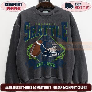 May include: A charcoal gray crewneck sweatshirt featuring a graphic print with the text SEATTLE FOOTBALL EST. 1976. The design includes a brown football and a dark blue football helmet centered within a diamond shape with green and blue accents.