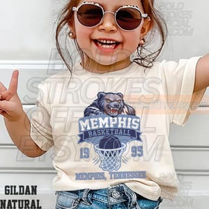 May include: A cream-colored t-shirt featuring a Memphis Basketball graphic. The design includes a bear, basketball, and the text "MEMPHIS BASKETBALL 1995 MEMPHIS, TENNESSEE". The shirt is made of Gildan Natural material.