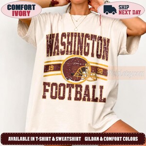 May include: An ivory short-sleeved t-shirt featuring a distressed graphic with the text WASHINGTON FOOTBALL in dark red. The design includes a dark red football helmet, the numbers 19 and 32, and horizontal gold stripes across the chest area.