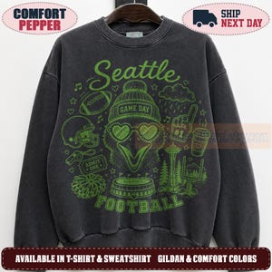 May include: A charcoal gray crewneck sweatshirt featuring a green graphic print of a bird wearing a beanie and heart-shaped glasses. The design includes the text Seattle Football, a football, a stadium, the Space Needle, and game day icons.