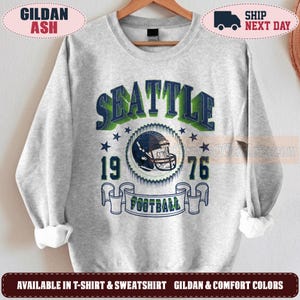 May include: A light gray crewneck sweatshirt featuring a graphic print with the text SEATTLE in dark blue and green, the numbers 19 and 76, a football helmet illustration, and a banner containing the word FOOTBALL in dark blue and green.