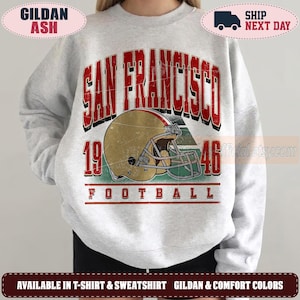 May include: A light grey crewneck sweatshirt featuring a distressed graphic print. The design includes the red text SAN FRANCISCO, a gold football helmet, the numbers 19 and 46, and the word FOOTBALL in red, with a vintage-style aesthetic.